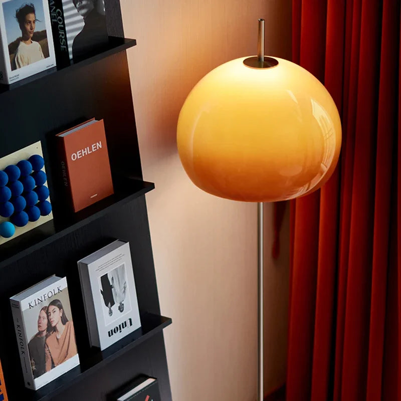 A modern floor lamp with a glowing gradient orange glass globe shade, standing next to a black ladder bookshelf in a warm, cosy room.