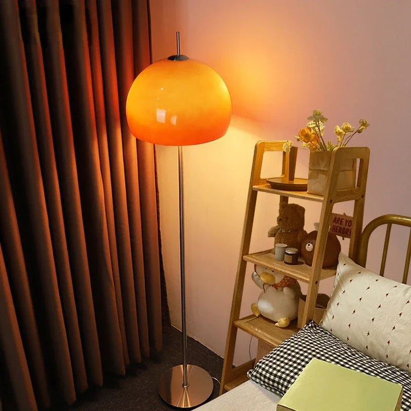 A mid-century modern floor lamp featuring a gradient orange glass shade glowing with warm light, set next to a wooden shelving unit in a cosy room.