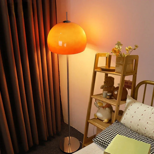A mid-century modern floor lamp featuring a gradient orange glass shade glowing with warm light, set next to a wooden shelving unit in a cosy room.