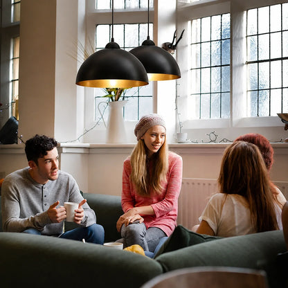 Two black and gold industrial dome pendant lights hanging above a group of friends chatting in a bright living room with large windows.