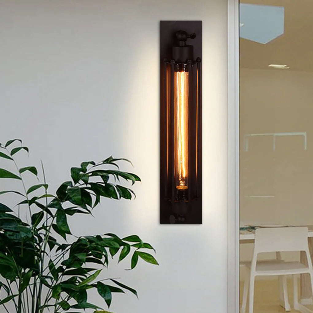 A vertical black industrial iron wall sconce with a glowing vintage filament bulb mounted on a white wall next to a green houseplant.