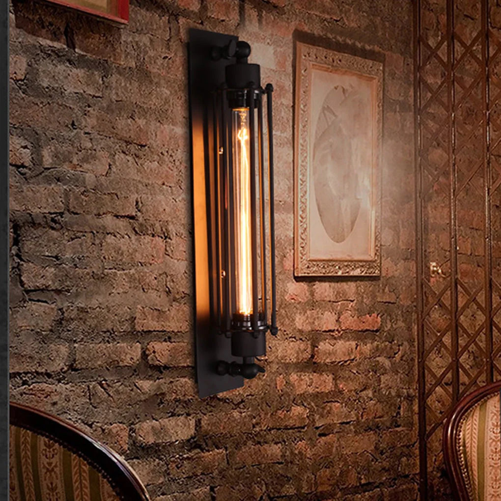 Black industrial iron wall sconce with a cylindrical cage design and a warm glowing vintage filament bulb mounted on a rustic exposed brick wall.