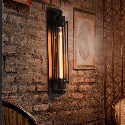 Black industrial iron wall sconce with a cylindrical cage design and a warm glowing vintage filament bulb mounted on a rustic exposed brick wall.