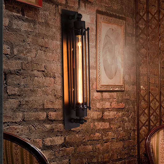 Black industrial iron wall sconce with a cylindrical cage design and a warm glowing vintage filament bulb mounted on a rustic exposed brick wall.