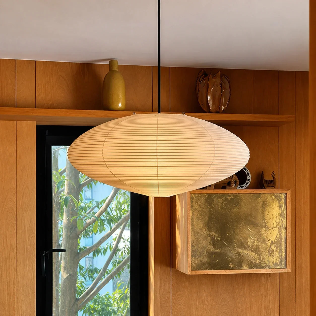 Japanese oval rice paper pendant light suspended in a minimalist interior with wood-panelled walls, a window view and decorative shelf ornaments.