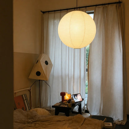 A large, glowing Japanese paper globe pendant light hangs in a minimalist bedroom, casting a soft, warm light over a low table and sheer curtains.