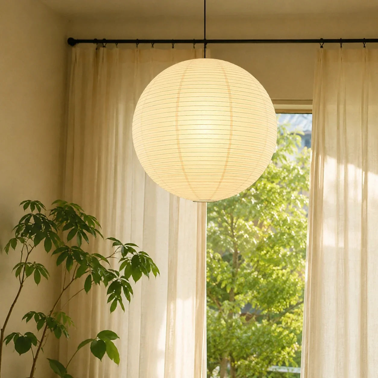 A minimalist Japanese paper globe pendant light hanging in a bright room with sheer curtains and a green houseplant, creating a warm Japandi aesthetic.