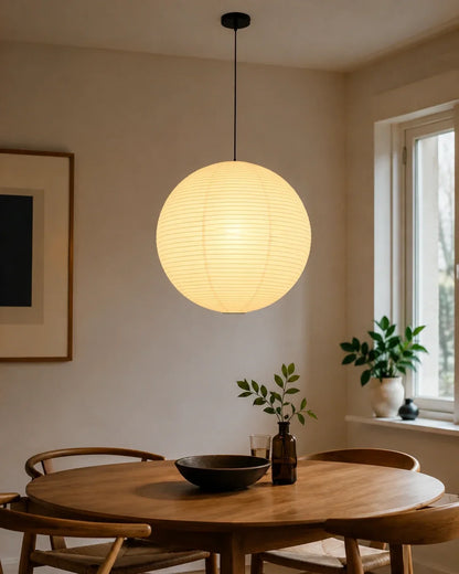 A minimalist Japanese paper globe pendant light hanging above a wooden dining table in a bright, Japandi-style room with soft, warm ambient lighting.