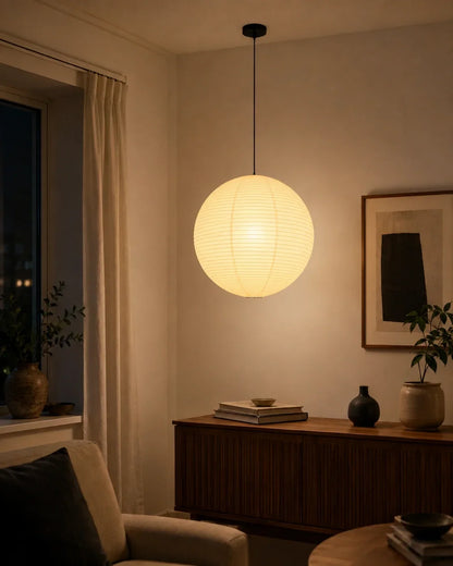 A glowing Japanese paper globe pendant light suspended in a minimalist living room, casting a warm, soft light over a wooden sideboard and decor.