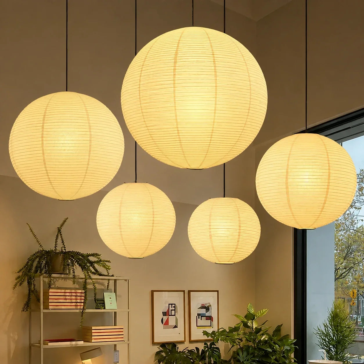 Five Japanese paper globe pendant lights of varying sizes suspended at different heights in a minimalist interior with indoor plants and wall art.