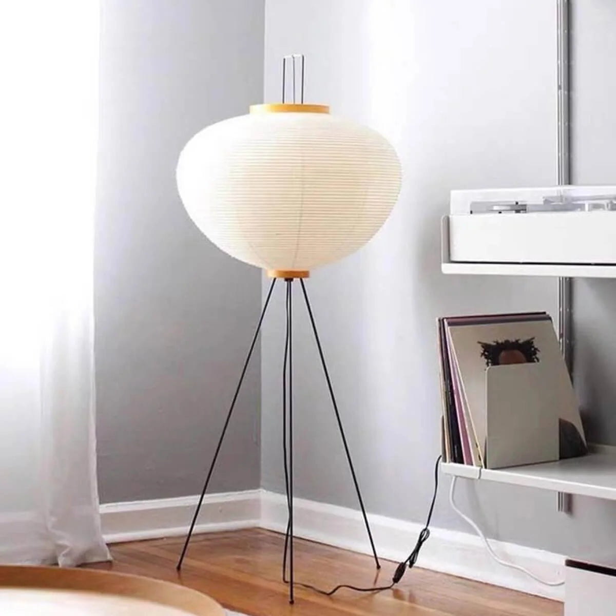 Japanese rice paper floor lamp with a white oval shade and black tripod base, standing on a wooden floor in a modern living room corner.