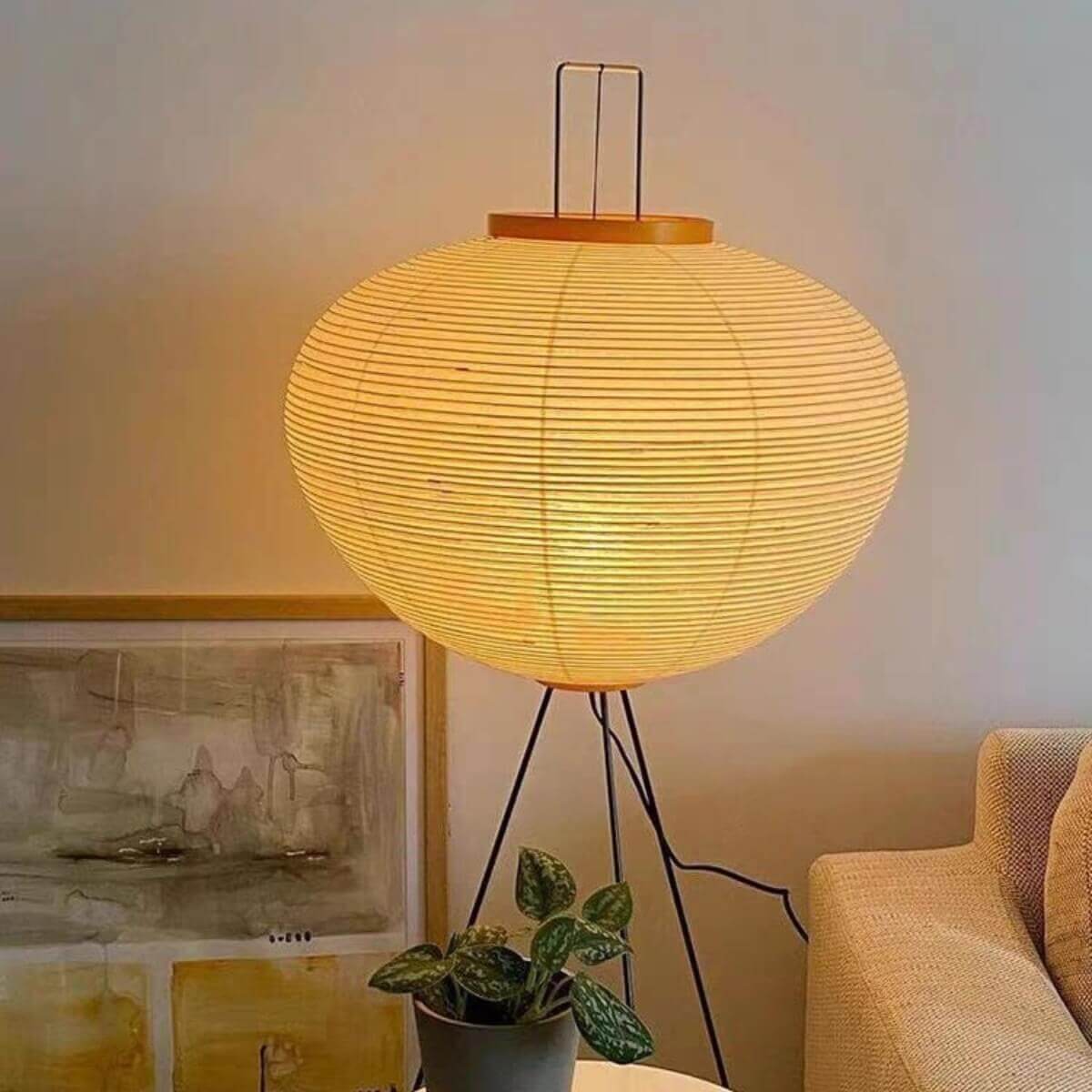 A glowing Japanese rice paper floor lamp with a black tripod base, emitting a warm light in a cosy living room next to a sofa.