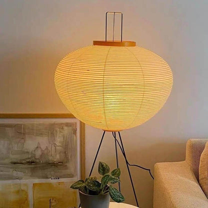 A glowing Japanese rice paper floor lamp with a black tripod base, emitting a warm light in a cosy living room next to a sofa.