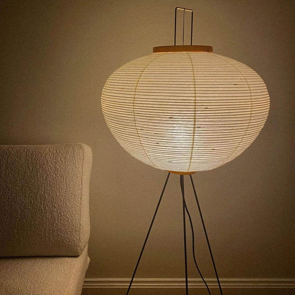 A Japanese rice paper floor lamp with a tripod base, glowing with warm light next to a textured beige armchair in a cosy living room.