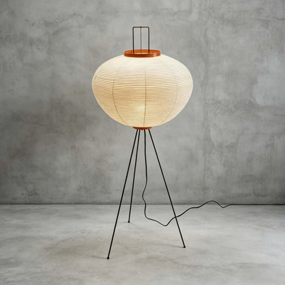 A lit Japanese rice paper floor lamp with a black tripod base, casting a warm glow against a minimalist grey concrete wall and floor.