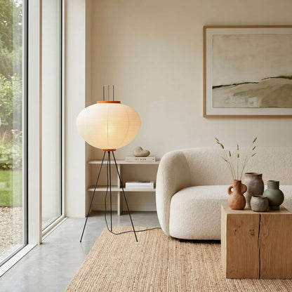 A Japanese rice paper floor lamp with a black tripod base glows warmly in a minimalist living room next to a white bouclé sofa and jute rug.