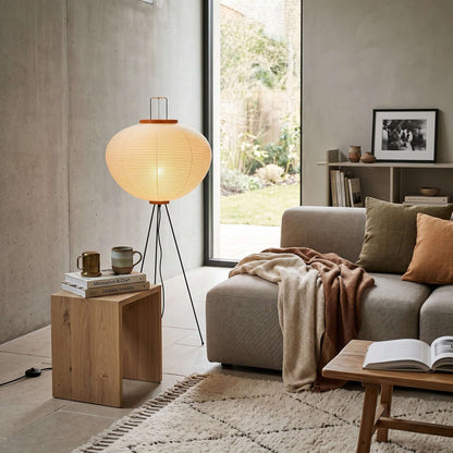 A Japanese rice paper floor lamp with a black tripod base glows warmly in a modern, cosy living room next to a grey sofa and wooden side table.