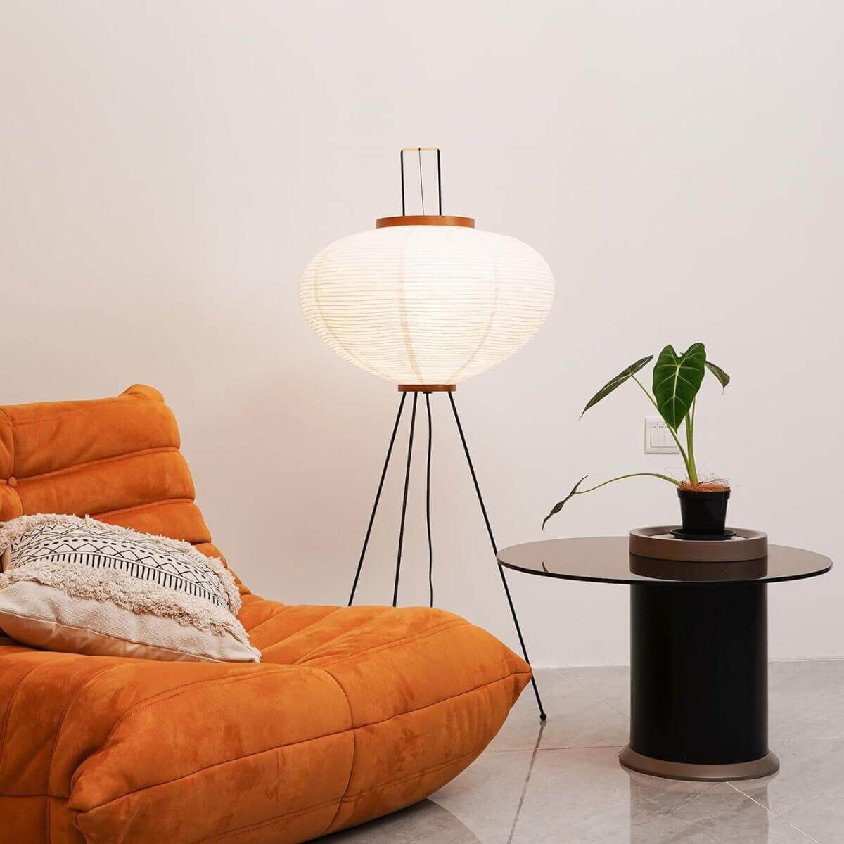 Japanese rice paper floor lamp with a black tripod base, illuminated next to an orange lounge chair and a glass side table in a living room.