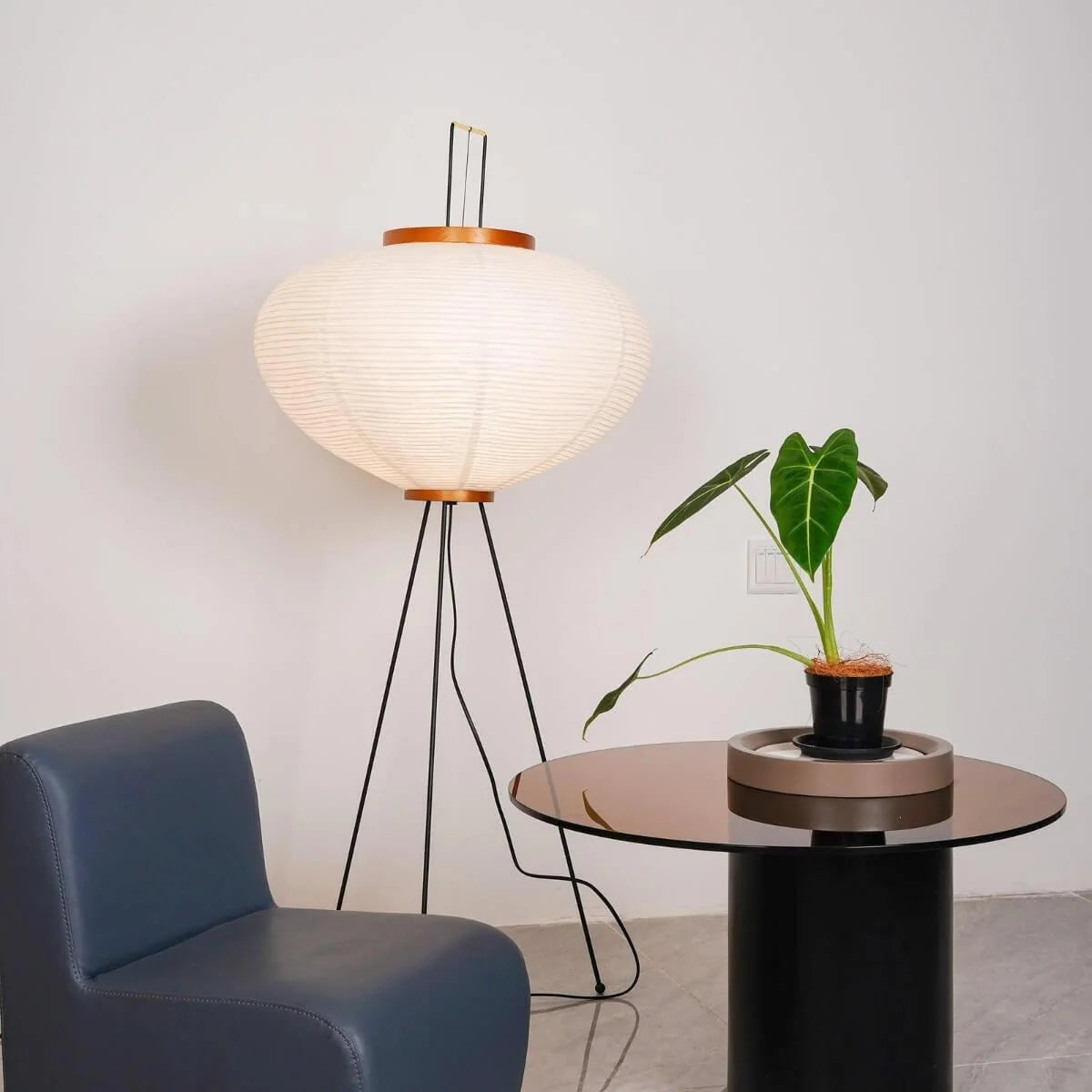 An illuminated Japanese rice paper floor lamp with a black tripod base, styled in a living room next to a blue armchair and a glass side table.