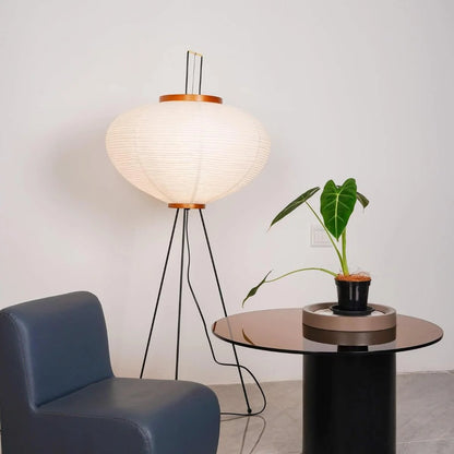 An illuminated Japanese rice paper floor lamp with a black tripod base, styled in a living room next to a blue armchair and a glass side table.