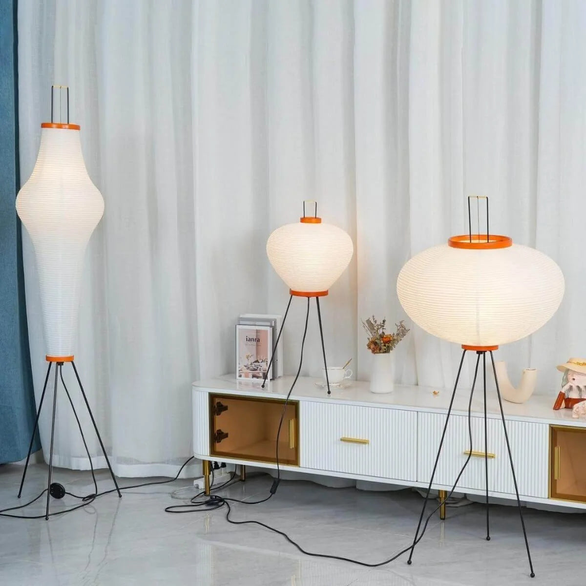 Three Japanese rice paper floor and table lamps with black tripod bases and orange rims, illuminated in a modern living room with white curtains.