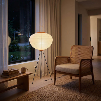 A Japanese rice paper floor lamp with a black tripod base glows warmly next to a wooden rattan armchair in a cosy living room setting.