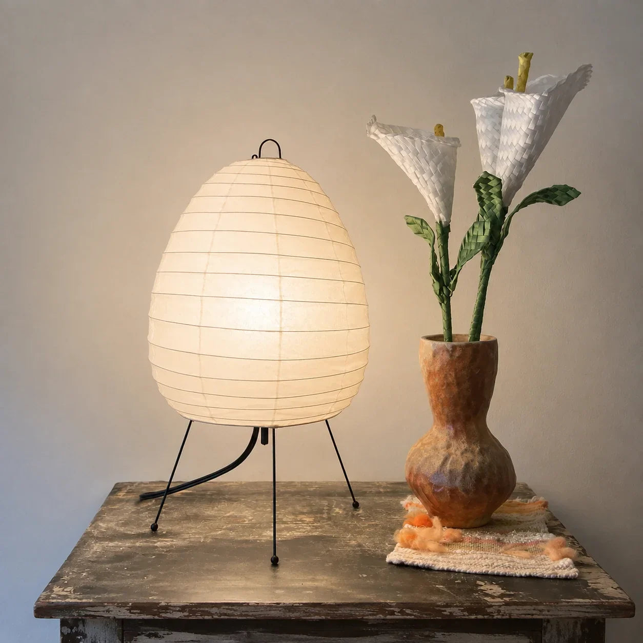 Japanese rice paper tripod table lamp emitting a warm glow, placed on a rustic wooden surface next to a textured vase with white decorative flowers.