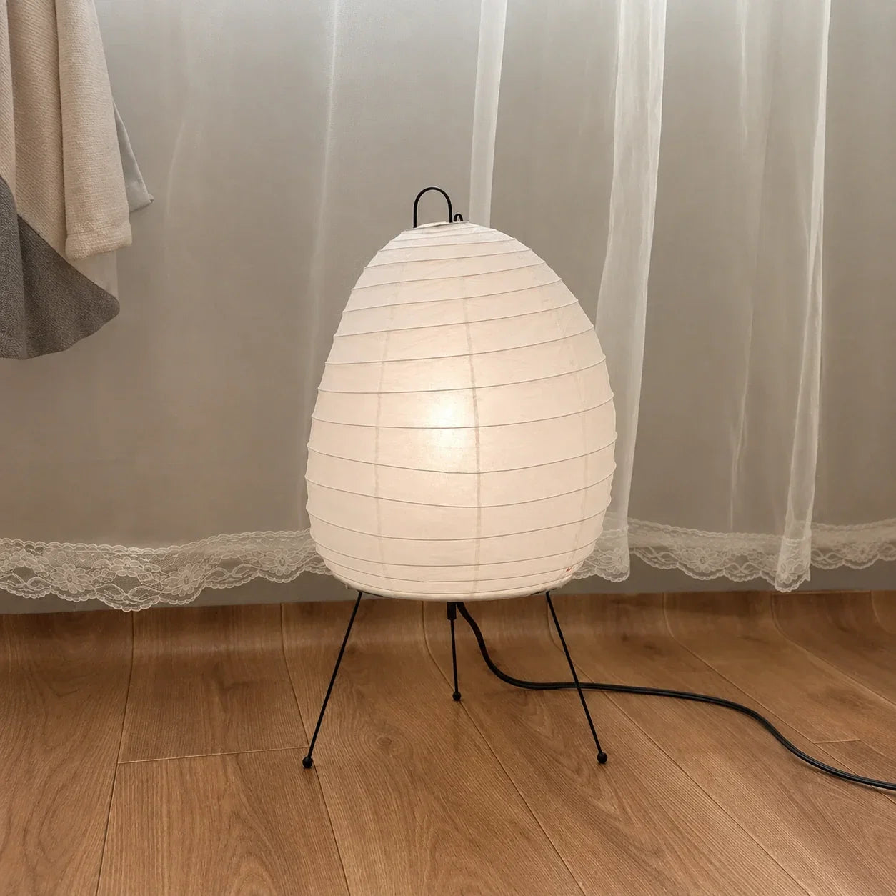 A Japanese-style rice paper table lamp with an oval shade and black tripod legs, glowing softly on a wooden floor in front of sheer white curtains.