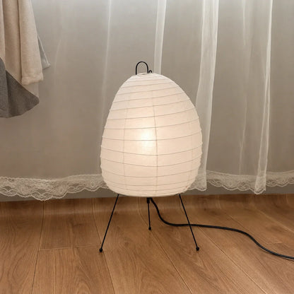 A Japanese-style rice paper table lamp with an oval shade and black tripod legs, glowing softly on a wooden floor in front of sheer white curtains.