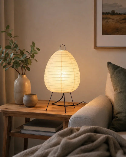 Japanese rice paper tripod table lamp emitting a warm, ambient glow on a wooden side table in a cosy Japandi-style bedroom interior.
