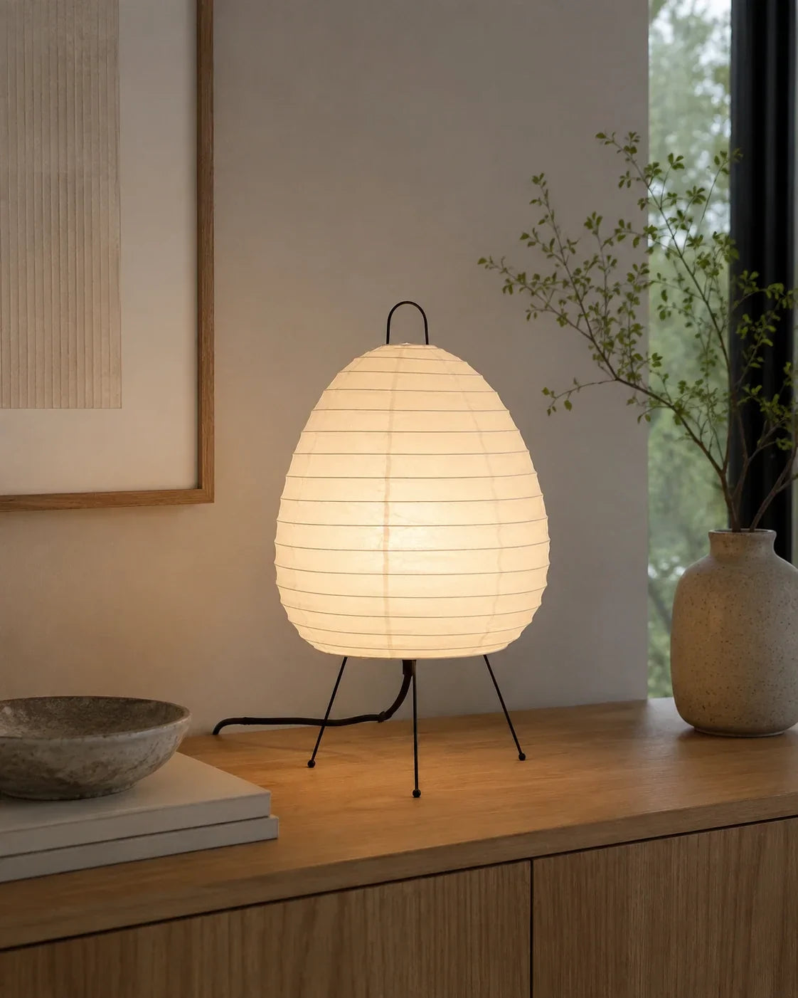 Japanese rice paper tripod table lamp with a warm glowing shade, resting on a wooden sideboard in a minimalist Japandi style living space.