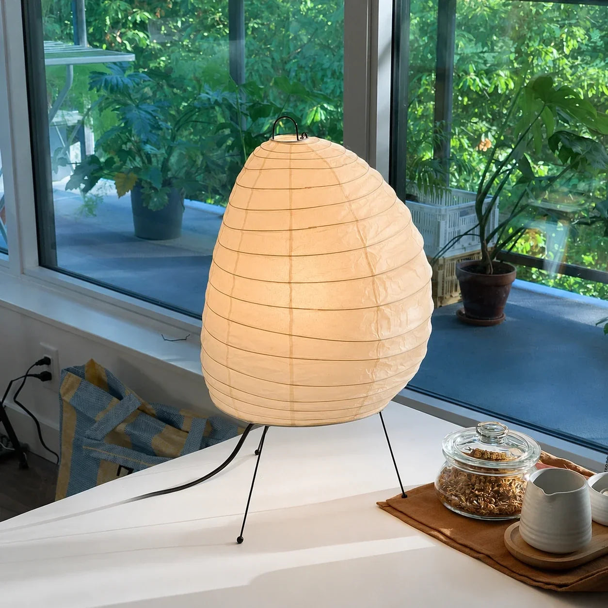 Japanese rice paper tripod table lamp with a warm glow, sitting on a white surface in front of a window with greenery, creating a Japandi aesthetic.