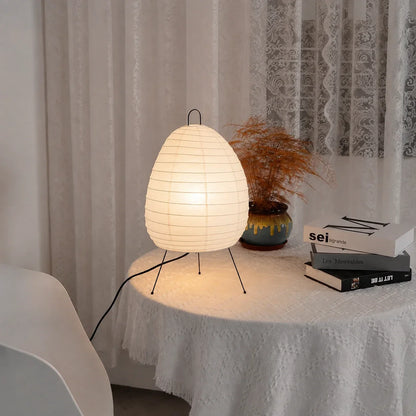 Japanese-style rice paper tripod table lamp emitting a warm glow on a lace-covered table next to a small potted plant and a stack of books.