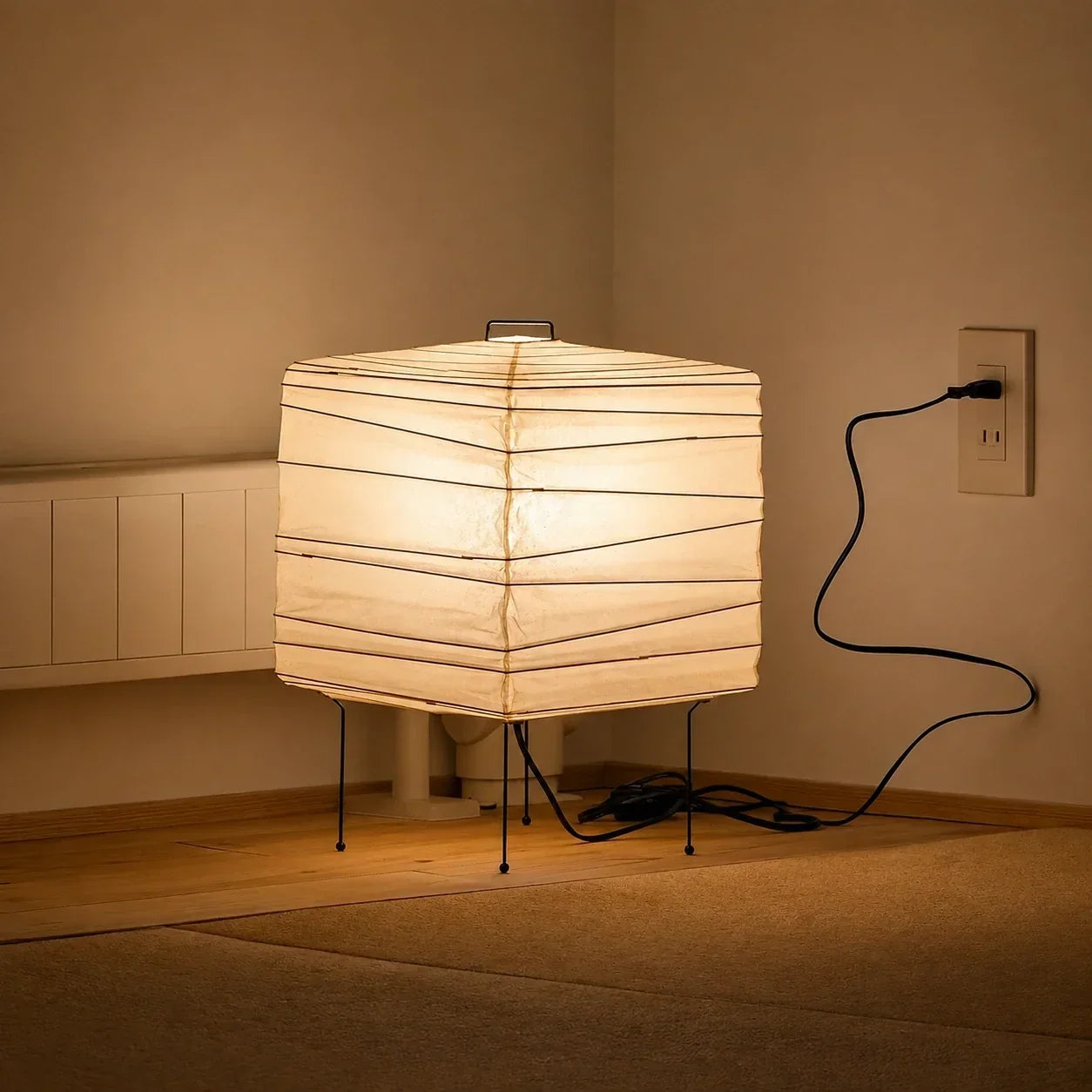 Minimalist square Japanese washi paper table lamp with a thin wire frame, emitting a warm glow on a wooden floor plugged into a wall socket.