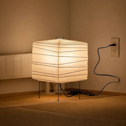 Minimalist square Japanese washi paper table lamp with a thin wire frame, emitting a warm glow on a wooden floor plugged into a wall socket.