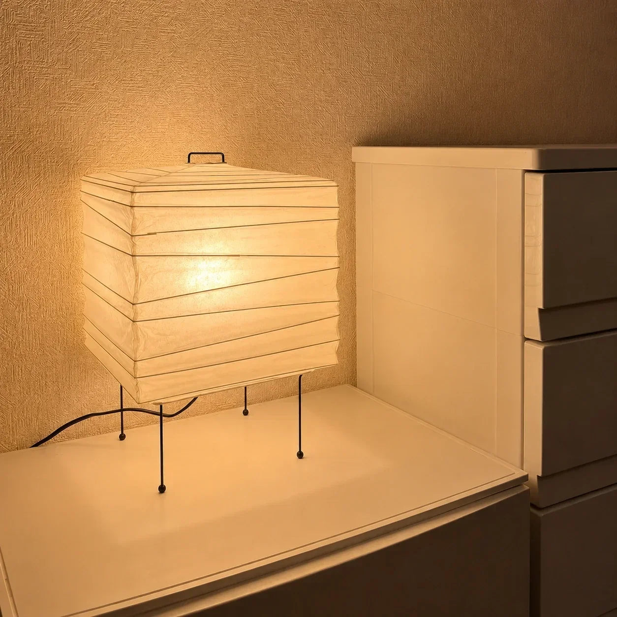 Minimalist square Japanese washi paper table lamp with a black wire frame, casting a warm, ambient glow on a white bedside unit in a bedroom setting.