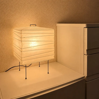 Minimalist square Japanese washi paper table lamp with a black wire frame, casting a warm, ambient glow on a white bedside unit in a bedroom setting.