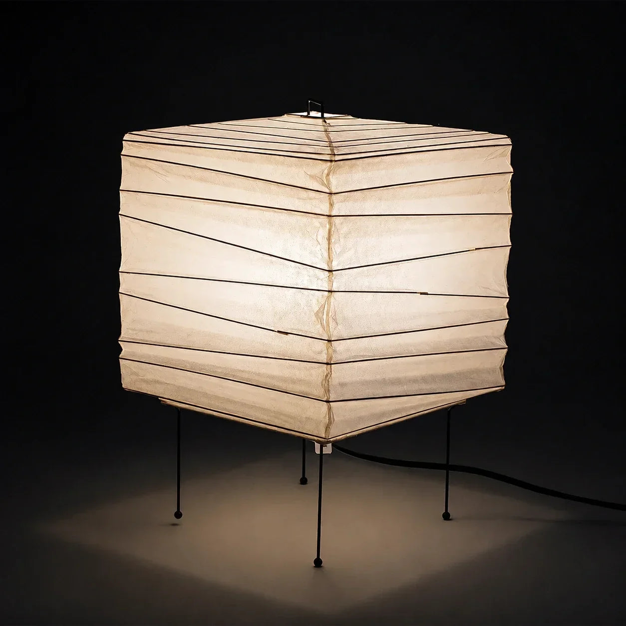 Minimalist Japanese square washi paper table lamp with a warm glow, supported by thin black metal legs against a dark background for ambient lighting.