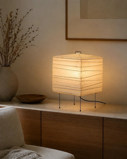 Square Japanese washi paper table lamp with a warm glow, set on a wooden sideboard in a minimalist Japandi living space with a vase of dried stems.