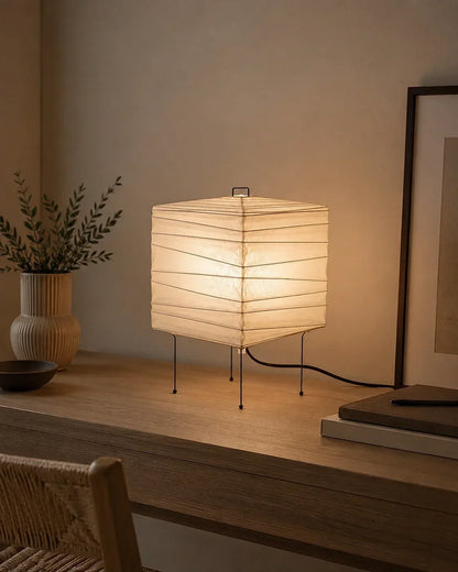 Square Japanese washi paper table lamp emitting a warm glow, set on a wooden desk in a minimalist Japandi-style room with a vase and framed artwork.
