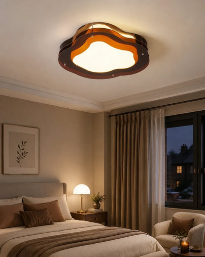 A modern, layered cloud-shaped LED ceiling light emitting a warm glow in a cosy bedroom with a bed, bedside lamp, and neutral decor.