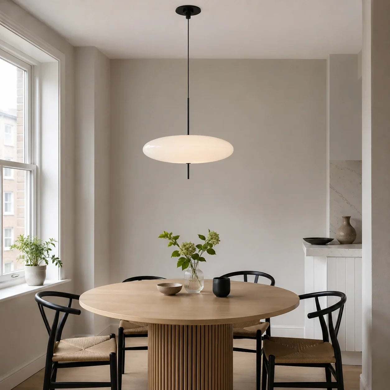 A modern flying saucer-shaped LED pendant light hanging above a round wooden dining table with black chairs in a minimalist dining room.