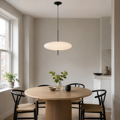 A modern flying saucer-shaped LED pendant light hanging above a round wooden dining table with black chairs in a minimalist dining room.