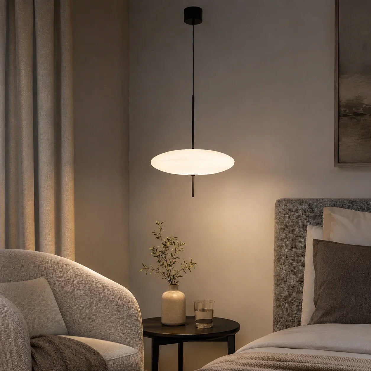 A modern LED pendant light with a glowing white flying saucer design, suspended above a bedside table in a cosy, minimalist bedroom.