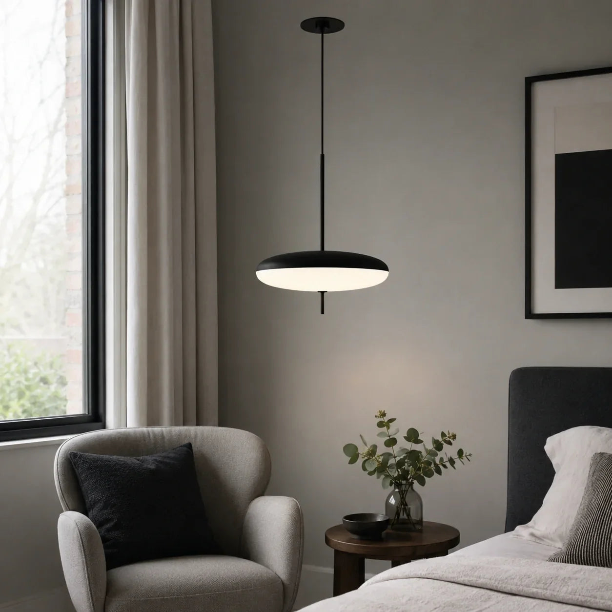 A modern black LED pendant light with a flying saucer design hanging in a minimalist bedroom next to a grey armchair and bedside table.