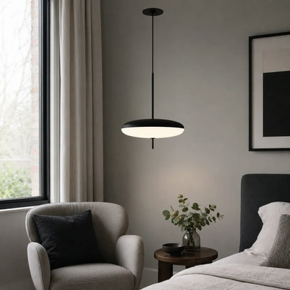 A modern black LED pendant light with a flying saucer design hanging in a minimalist bedroom next to a grey armchair and bedside table.