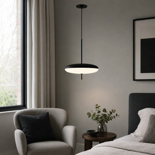 A modern black LED pendant light with a flying saucer design hanging in a minimalist bedroom next to a grey armchair and bedside table.