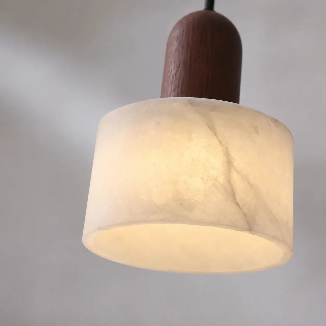 Minimalist pendant light featuring a cylindrical white marble shade with natural veining and a warm wooden top detail, glowing against a neutral wall.