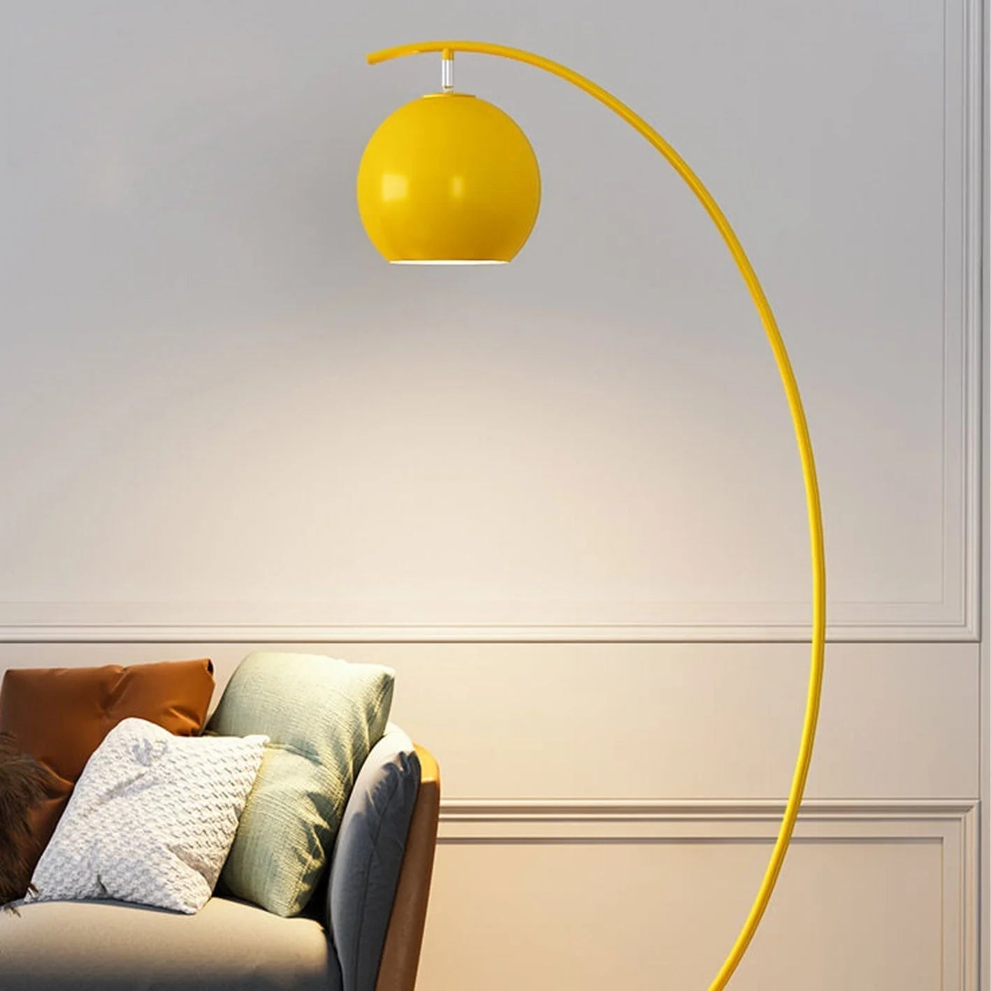 A minimalist yellow LED arc floor lamp with a spherical shade casting a warm glow over a grey sofa with cushions in a modern living room.