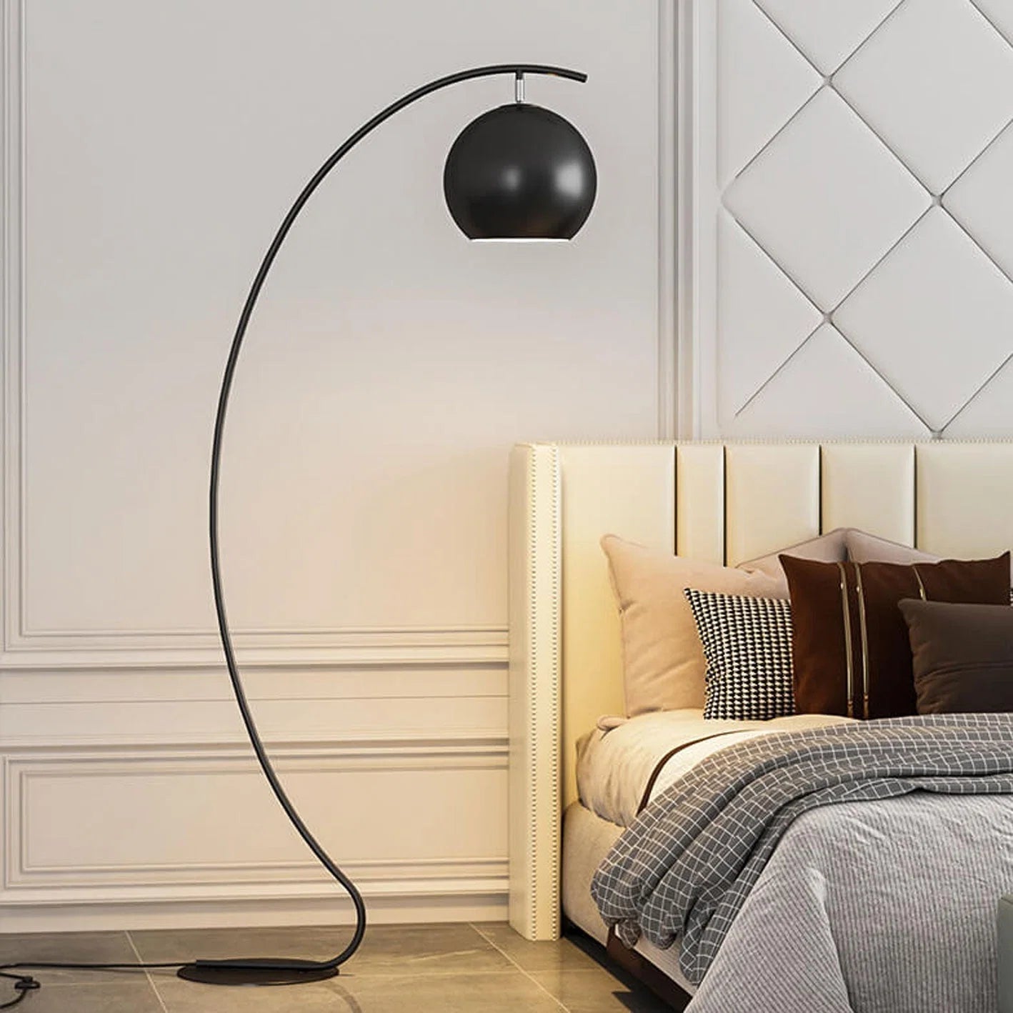 A minimalist black LED arc floor lamp with a spherical shade standing next to a bed with a cream headboard in a modern bedroom.